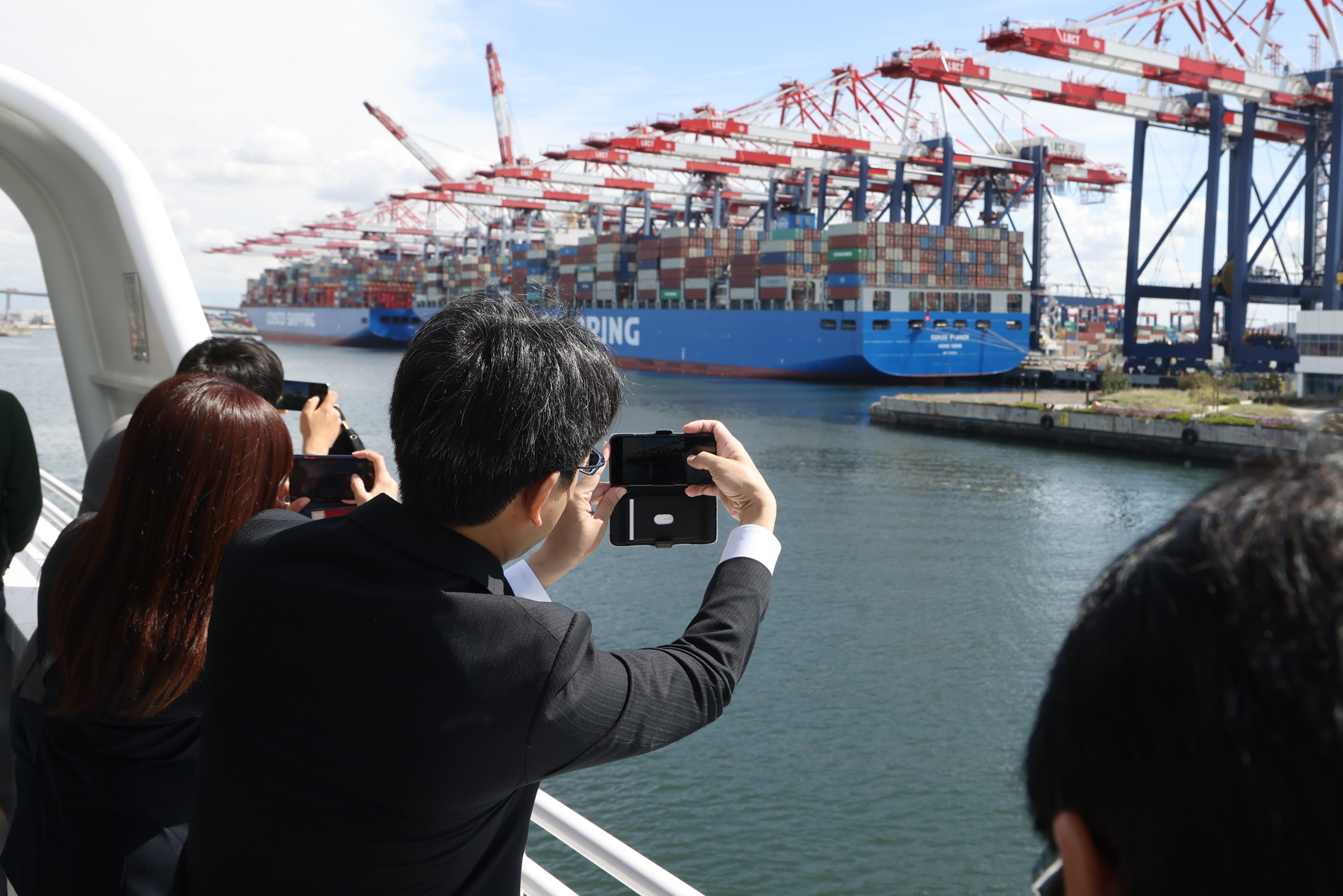A photo of 3 people taking pictures of large container ship and drayage equipment on the upper deck of a boat