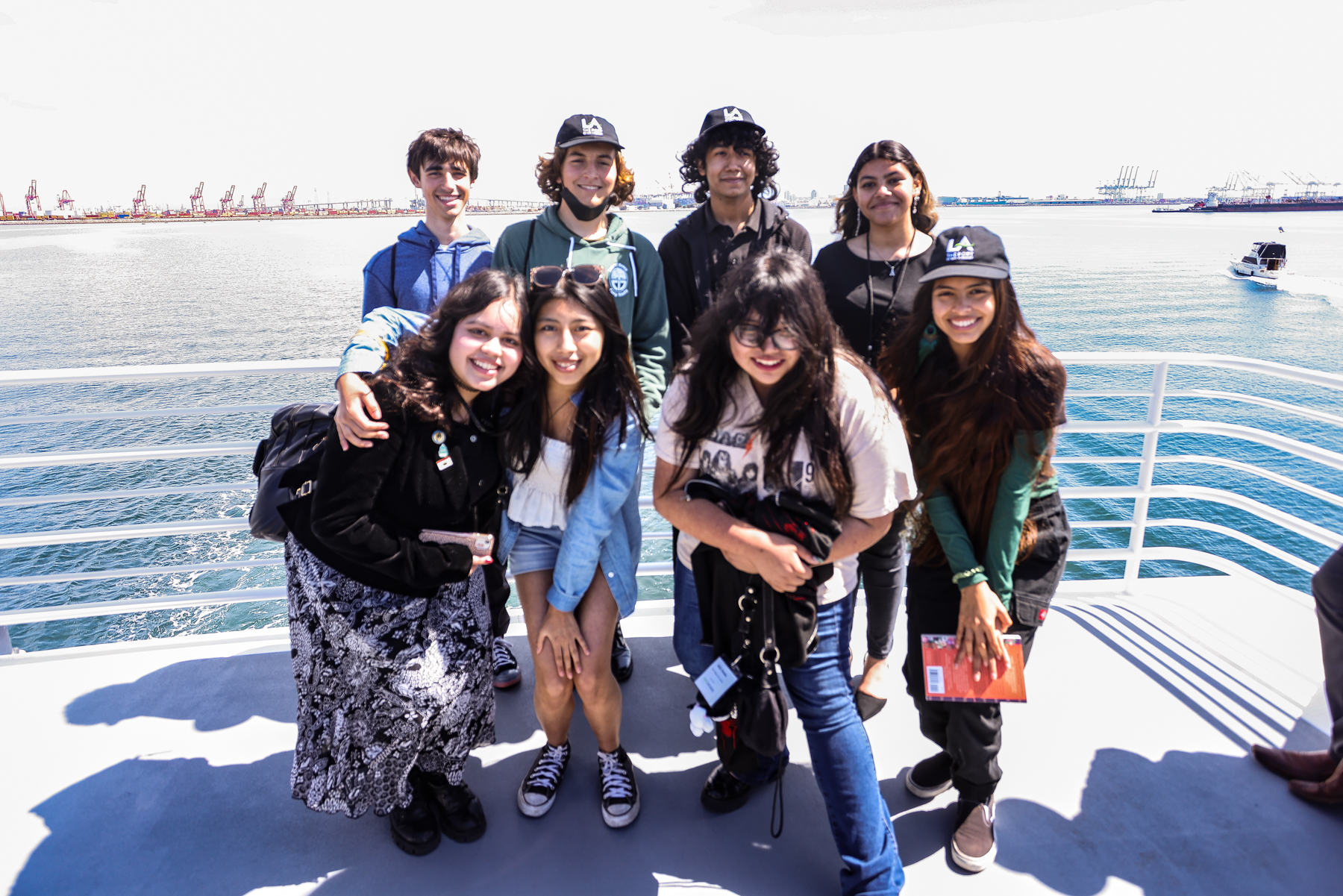 A photo of 8 students on the upper deck of a boat with San Pedro Bay and port drayage equipment in the background